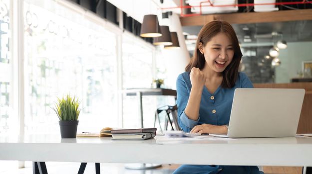 young Asian businesswomen show joyful expression of success at work smiling happily with a laptop computer in a modern office