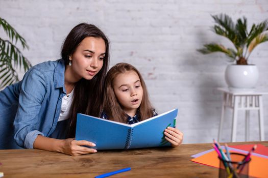 Mom helps my daughter do her homework in the kitchen. Mom and daughter are engaged in reading. They are in a good mood and smile.