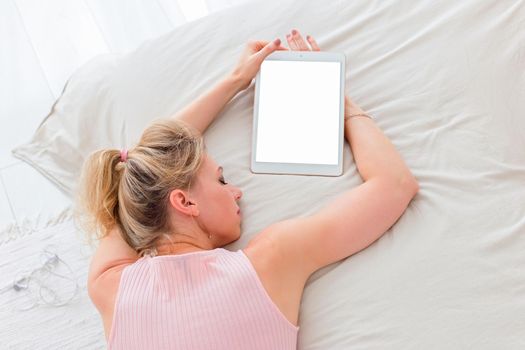 Tired slender woman with blond hair, pink top lies in the morning on a white bed, a digital tablet with a white screen lies nearby, top view. Mock up. Copy space