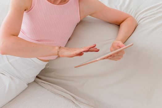 A slender woman a pink top and white pants, sits on the floor in the morning, near a white bed, look at digital tablet, scrolls screen of device. Copy space