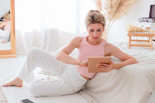 A beautiful slender woman with blond hair, a pink top and white pants, sits on the floor in the morning, near a white bed, look at digital tablet, a smartphone lies nearby. Copy space