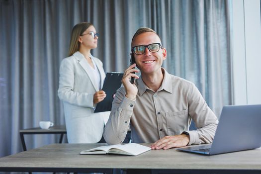 A male manager is talking on a mobile phone while sitting at a table with a computer and a notepad. Working atmosphere in an office with large windows
