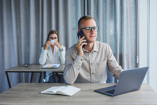 A male manager is talking on a mobile phone while sitting at a table with a computer and a notepad. Working atmosphere in an office with large windows