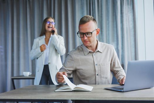 A business woman in front of a male colleague who works at a laptop. Business cooperation and teamwork. European office workers at the table in the office. Modern successful people