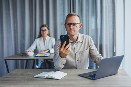 A male manager is talking on a mobile phone while sitting at a table with a computer and a notepad. Working atmosphere in an office with large windows