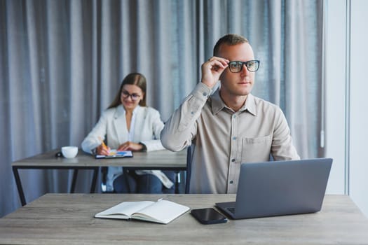 Portrait of a cheerful man, IT professional, working with a modern laptop, sitting at a table and smiling at the camera during a break, a happy human programmer in vision correction glasses