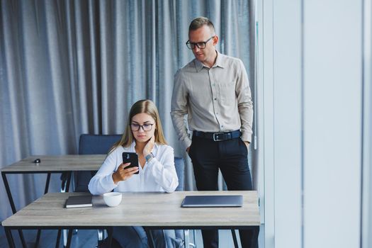 Confident male and female colleagues in elegant casual wear are discussing a project in the workplace, enjoying corporate friendship, happy partners are talking about office networks on a mobile phone