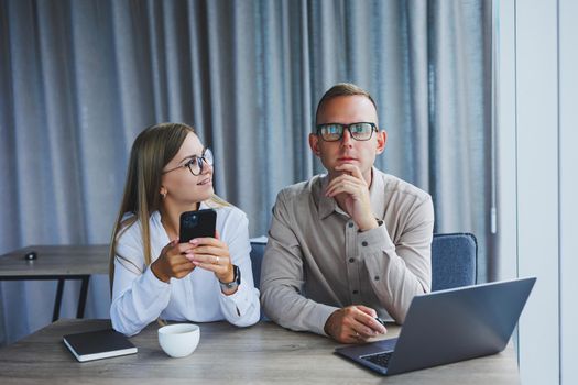 Cheerful businesspeople using a laptop in an office. Happy young entrepreneurs smiling while working together in a modern workspace. Two young businesspeople sitting together at a table.