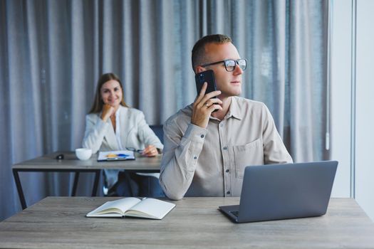 A male manager is talking on a mobile phone while sitting at a table with a computer and a notepad. Working atmosphere in an office with large windows