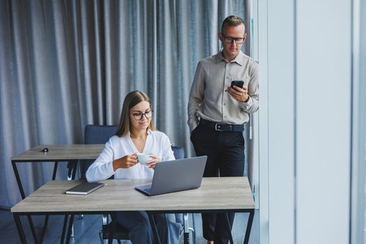 Confident male and female colleagues in elegant casual wear are discussing a project in the workplace, enjoying corporate friendship, happy partners are talking about office networks on a mobile phone