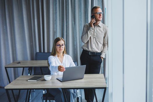 Confident male and female colleagues in elegant casual wear are discussing a project in the workplace, enjoying corporate friendship, happy partners are talking about office networks on a mobile phone