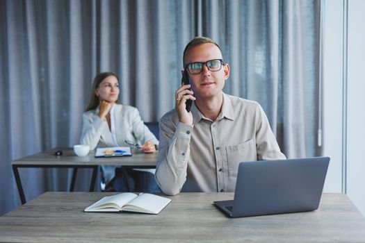 A male manager is talking on a mobile phone while sitting at a table with a computer and a notepad. Working atmosphere in an office with large windows