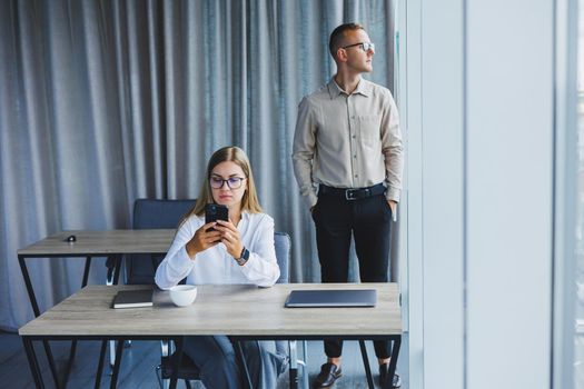 Confident male and female colleagues in elegant casual wear are discussing a project in the workplace, enjoying corporate friendship, happy partners are talking about office networks on a mobile phone