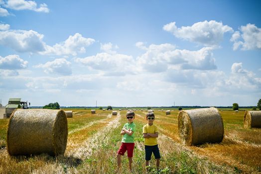 Two little boy stand among round haystack. Field with round bales after harvest under blue sky. Big round bales of straw, sheaves, haystacks.