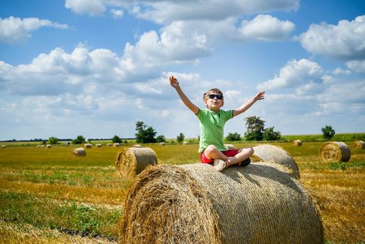 Little boy sits on a round haystack relaxed with hands up. Field with round bales after harvest under blue sky. Big round bales of straw, sheaves, haystacks.