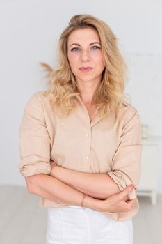 Portrait of a beautiful woman, 30-40 years old, with blond hair, stands in a beige linen shirt and white trousers, in a white room interior. Vertical