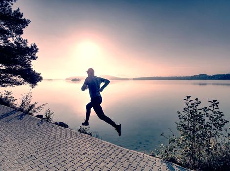 Freeze sportsman in  running pose.  Runner is sprinting along mountain lake within morning moody atmosphere.