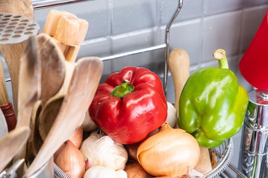 small basket in the kitchen next to the utensils with garlic, onions, shallots and red yellow and green peppers.