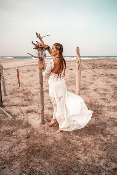 Model in boho style in a white long dress and silver jewelry on the beach. Her hair is braided, and there are many bracelets on her arms