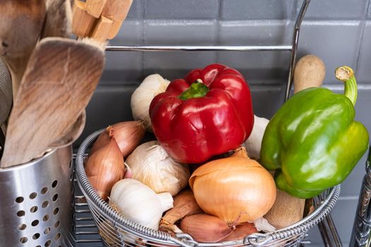 small basket in the kitchen next to the utensils with garlic, onions, shallots and red yellow and green peppers.