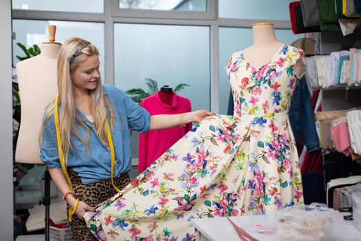A clothing designer hangs a floral dress on a mannequin and checks how the whole thing looks. A blonde woman in the back of a tailor's workshop.