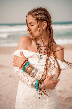 Model in boho style in a white long dress and silver jewelry on the beach. Her hair is braided, and there are many bracelets on her arms