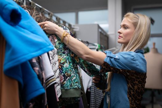 A woman looks at the new clothes hanging on the hangers. A blonde woman with long hair looks for the right blouse for her.