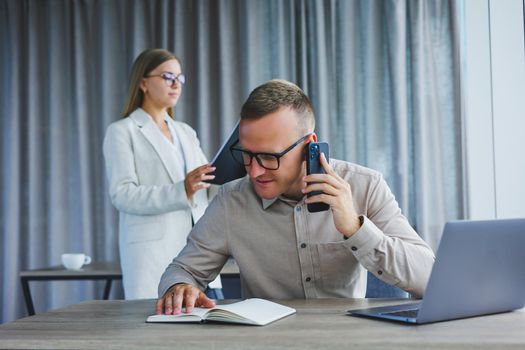 A male manager is talking on a mobile phone while sitting at a table with a computer and a notepad. Working atmosphere in an office with large windows
