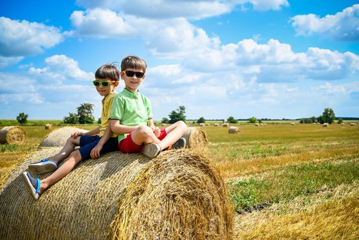 Two little brother boys sits on a round haystack relaxed. Field with round bales after harvest under blue sky. Big round bales of straw, sheaves, haystacks. Agriculture concept.