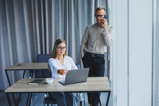 Confident male and female colleagues in elegant casual wear are discussing a project in the workplace, enjoying corporate friendship, happy partners are talking about office networks on a mobile phone