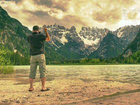 Photo traveler is taking memory photo of lake scenery. Blue lake between high mountains, peaks touch in blue sky. Travel lifestyle concept