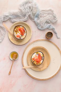 Yogurt with grapefruit, granola, chia and honey served in half a coconut shell on a rose marble kitchen countertop.