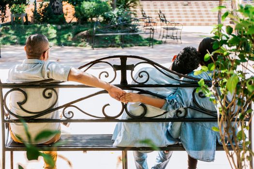 Rear view of unfaithful woman holding hands with another man while boyfriend hugs her. Unfaithful couple holding hands on a park bench. Cheating girlfriend with her boyfriend holding another man hand