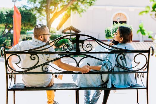 Cheating girlfriend with her boyfriend holding another man hand in a park. Unfaithful woman sitting holding hands with another man while boyfriend hugs her. love triangle concept