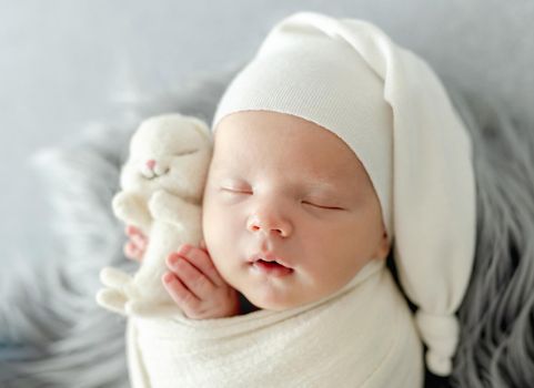 Newborn baby child swaddled in fabric sleeping and holding bunny toy. Sweet infant kid napping on fur