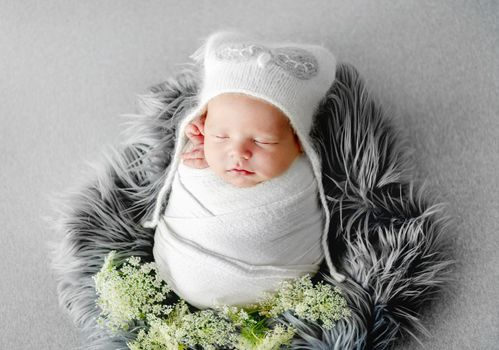 Newborn baby child swaddled in white fabric sleeping. Sweet infant kid wearing hat napping on fur