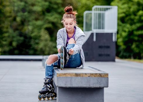 Cute girl roller skater sitting in city park and smiling. Pretty female teenager posing during rollerskating