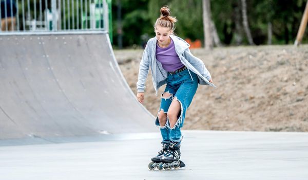 Beautiful girl roller skater riding in city park with ramp. Pretty female teenager rollerskating in casual clothes