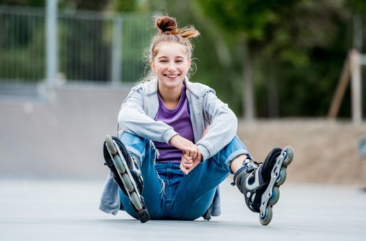 Cute girl roller skater sitting on ground in city park and smiling. Pretty female teenager with hairstyle posing during rollerskating