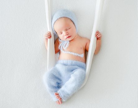 Newborn baby child sleeping on fabric swing. Sweet infant kid wearing knitted pants and hat napping
