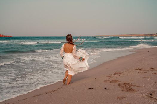 Model in boho style in a white long dress and silver jewelry on the beach. Her hair is braided, and there are many bracelets on her arms