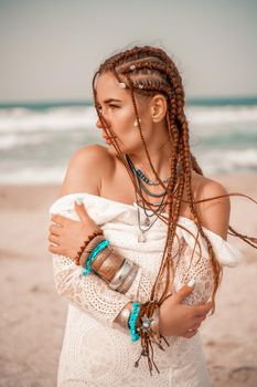 Model in boho style in a white long dress and silver jewelry on the beach. Her hair is braided, and there are many bracelets on her arms