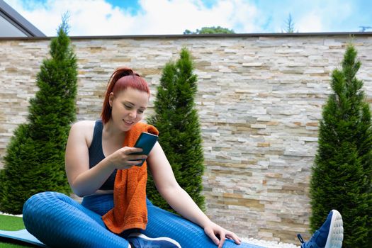 Attractive young fitness girl looking at her smart phone sitting on a pilates ball outdoors. girl exercising while sharing it on her social networks. health and wellness concept. natural light in the garden.