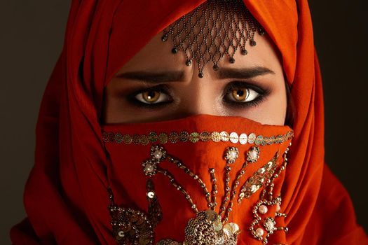 Close-up portrait of a moody young girl with charming smoky eyes and jewelry on the forehead, wearing the terracotta hijab decorated with sequins. She is posing and looking at the camera on a dark background. Human emotions, facial expression concept. Trendy colors. Arabic style.