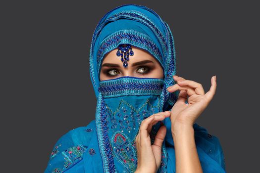 Close-up portrait of a nice young woman with beautiful smoky eyes wearing a blue hijab decorated with sequins and jewelry. She is gesticulating and looking away on a dark background. Human emotions, facial expression concept. Trendy colors. Arabic style.