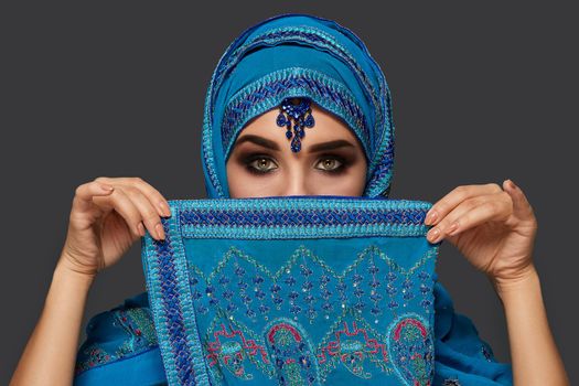 Close-up portrait of a young female with smoky eyes wearing an elegant blue hijab decorated with sequins and jewelry. She is covering her face with a shawl and looking at the camera on a dark background. Human emotions, facial expression concept. Trendy colors. Arabic style.