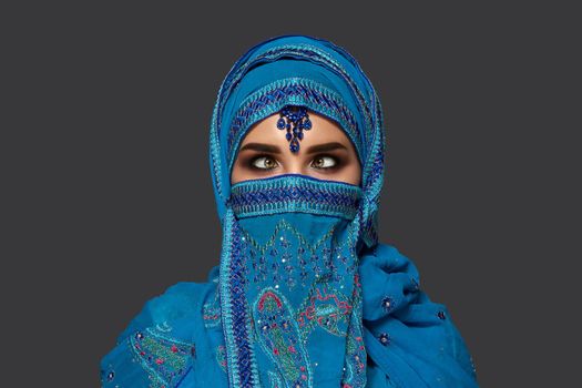 Close-up portrait of a beautiful girl with charming smoky eyes wearing a blue hijab decorated with sequins and jewelry. She is posing and making faces on a dark background. Human emotions, facial expression concept. Trendy colors. Arabic style.