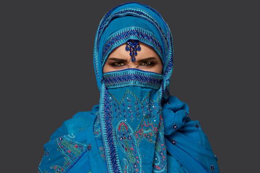 Close-up portrait of a nice girl with charming smoky eyes wearing an elegant blue hijab decorated with sequins and jewelry. She is posing and making faces on a dark background. Human emotions, facial expression concept. Trendy colors. Arabic style.