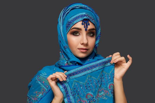 Close-up portrait of arabic girl with smoky eyes wearing a blue hijab decorated with sequins and jewelry. She has opened her face and looking at the camera on a dark background. Human emotions, facial expression concept. Trendy colors. Arabic style.