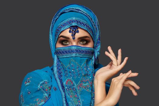 Close-up portrait of a pretty girl with charming smoky eyes wearing a blue hijab decorated with sequins and jewelry. She is gesticulating and looking at the camera on a dark background. Human emotions, facial expression concept. Trendy colors. Arabic style.